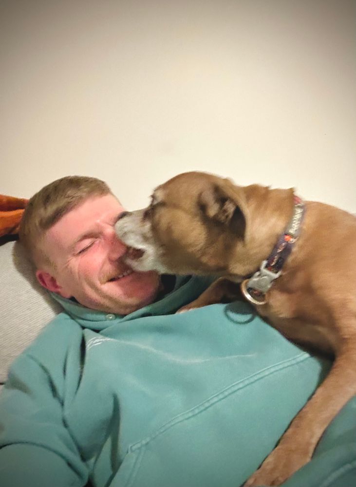 Selfie of a smiling man in a green hoodie with a Staffordshire Terrier licking his face abruptly while he was taking the picture.