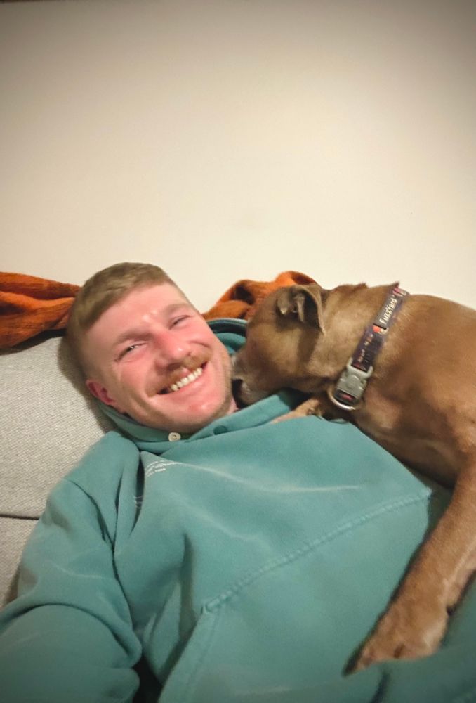 Selfie of a smiling man in a green hoodie with a Staffordshire Terrier licking his neck
