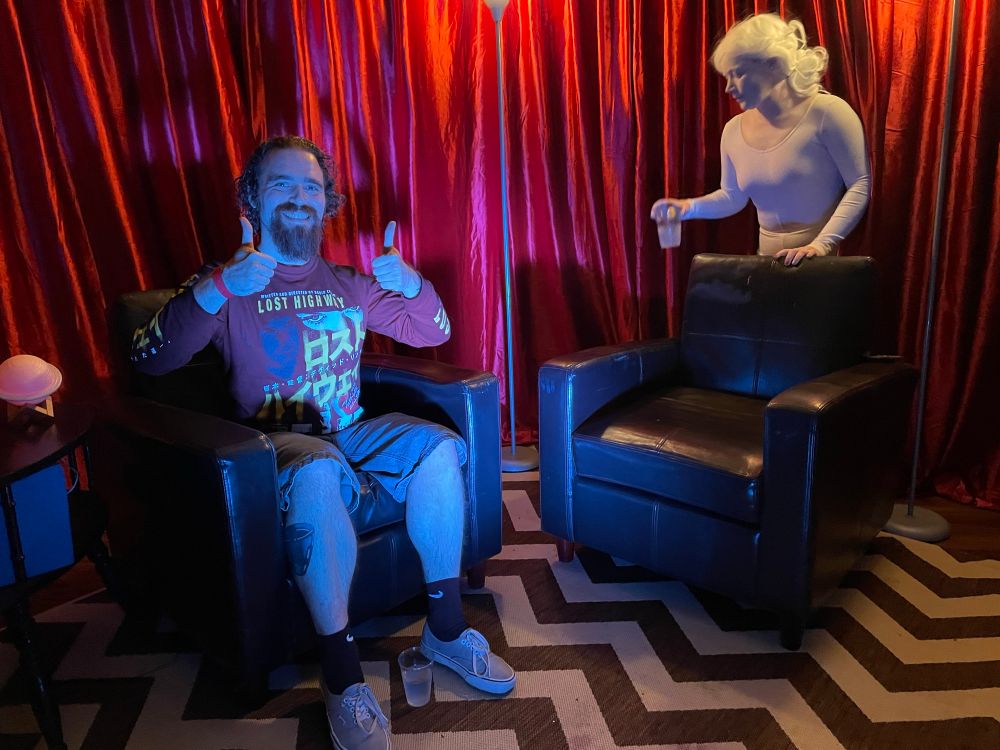 Man giving double thumbs up in a mock up of the Red Room from “Twin Peaks.” There’s a living statue on the other side of the room.