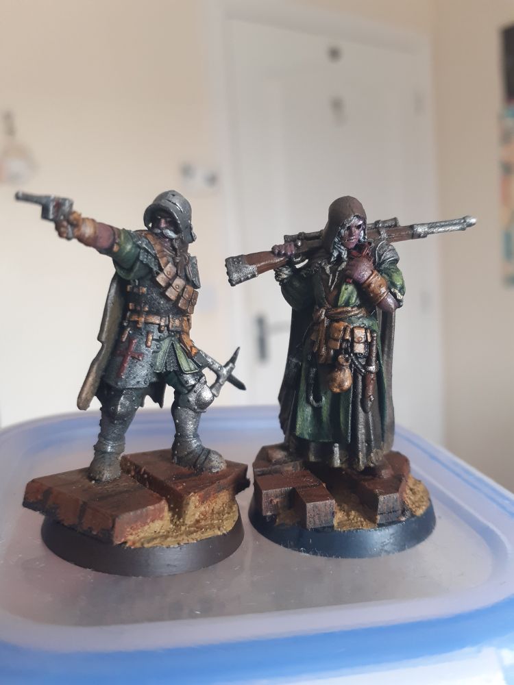 Image is of a pair of soldiers in armour that mixes elements of ww1 infantry and medieval knights. The man on the left wields a pistol and warhammer while sporting a prominent moustache. The woman on the right is an eyeless sniper.