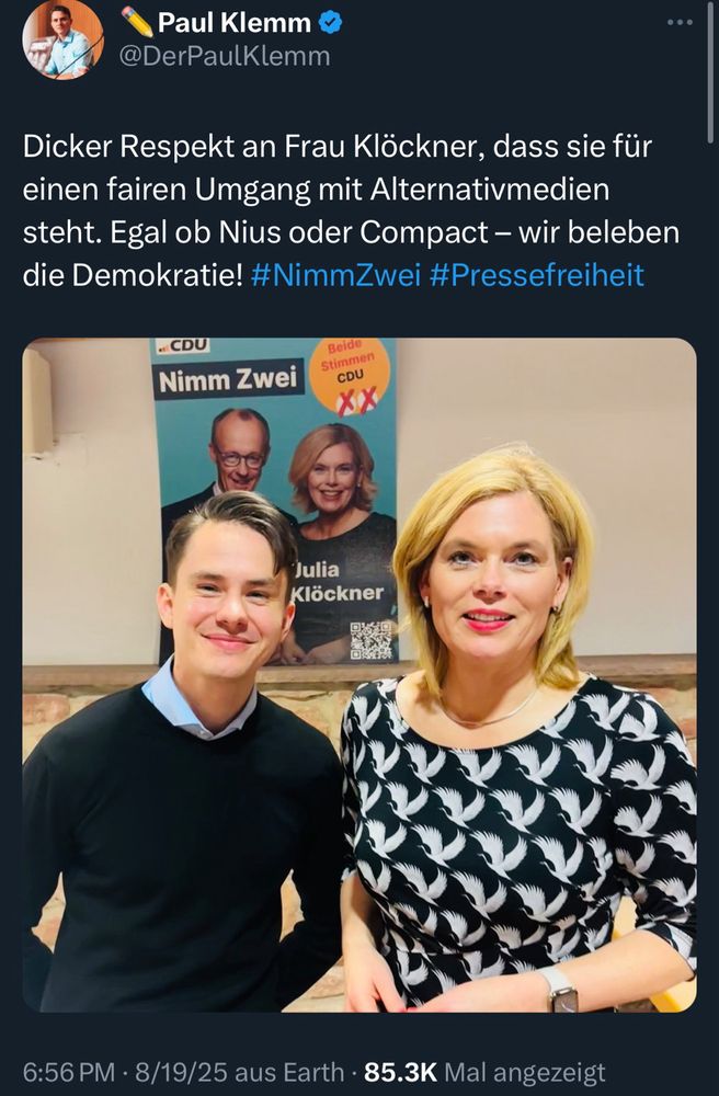 Tweet: Paul Klemm in Hemd und Pulli mit Klöckner in schwarzem Kleid mit weißen Kranichen vor Wahlplakat. 