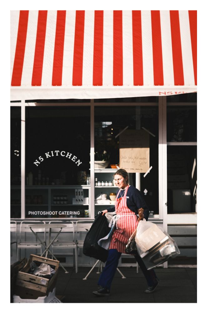 A chef in a red and white striped apron taking out bags of rubbish and recycling in from of a restaurant (N5 Kitchen) with a red and white striped awning. She is squinting in a ray of light