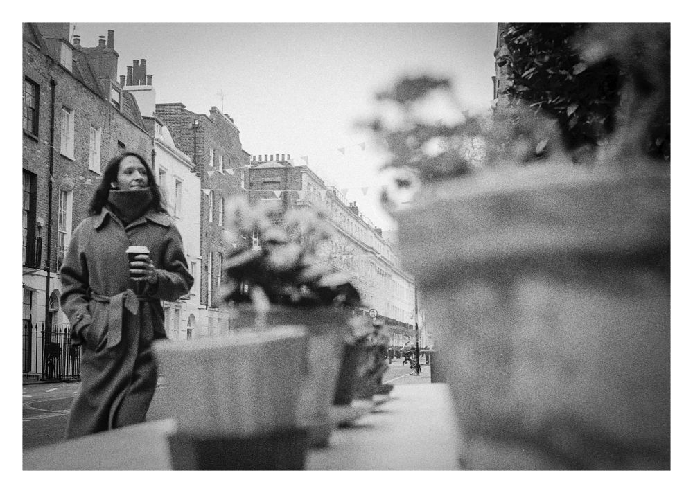 A lady in a long wool coat and big scarf carrying a coffee cup walking down a terraced street past a table of pot plants. Black and white. 