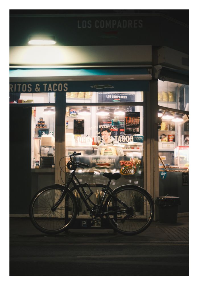A bicycle chained up in front of a burrito and taco restaurant (LOS CAMPADRES) at night. A chef is visible through the window.