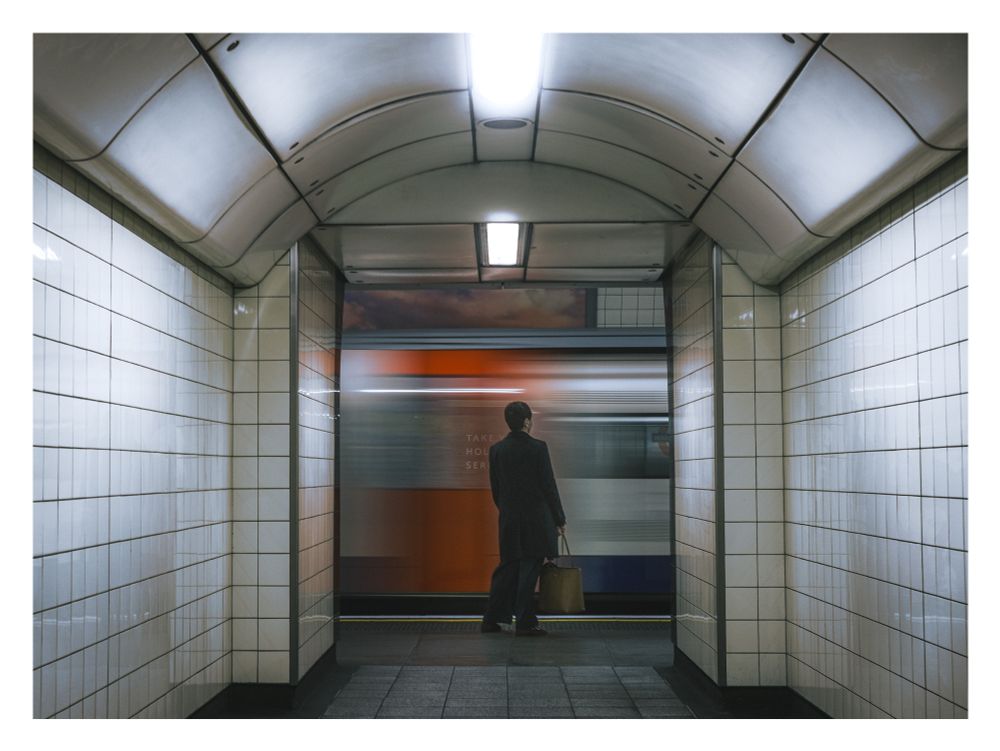 View through a tiled tunnel between tube platforms. A person holding a bag is at the end of the tunnel facing towards a motion-blurred train that is arriving. 