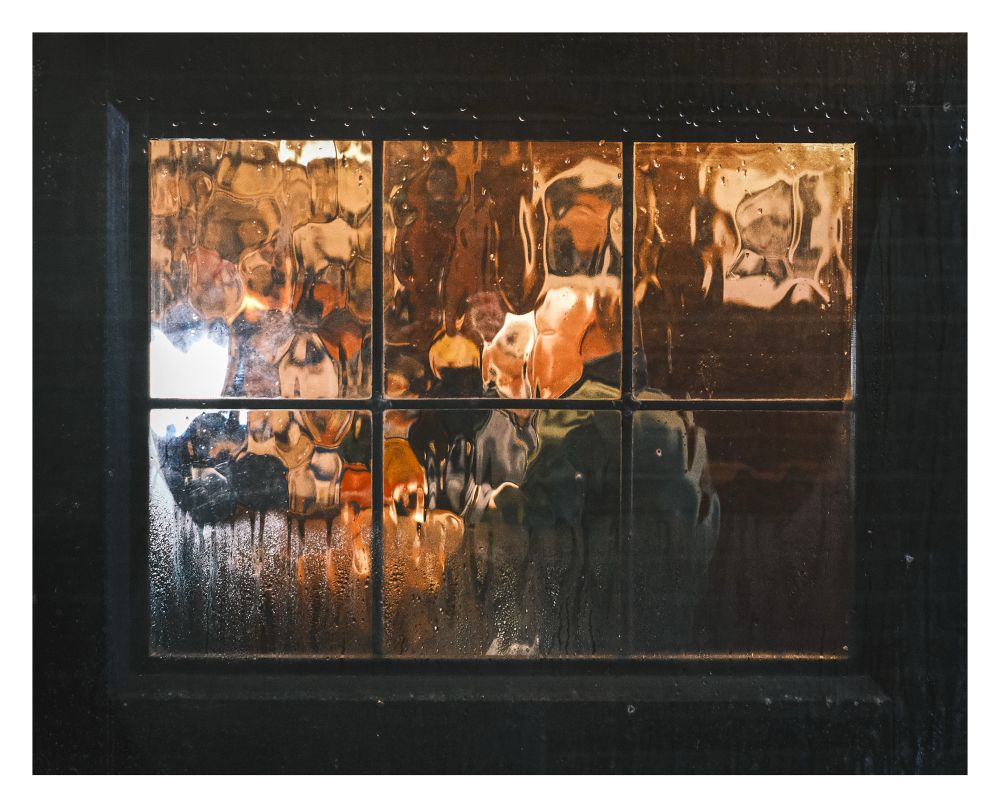 A figure seen having a drink (maybe) through a six-pane mottled pub window with some condensation on it at night.