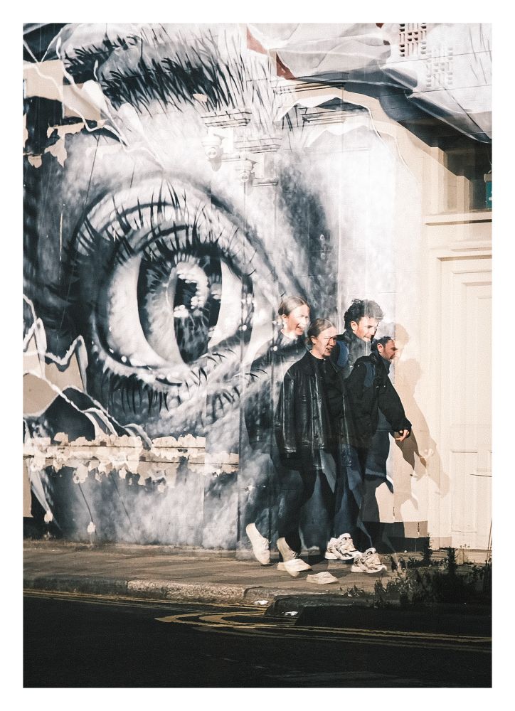 Two people walking in front of a huge street art eye drawn on a wall. They’re seen through a reflection which makes it look like there is two of everything/everyone slightly offset