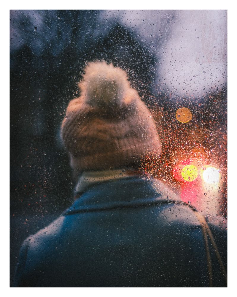Head and shoulders of someone in a bobble hat seen from behind through a rain drop covered bus shelter window. There’s low light and some oncoming traffic creating bokeh in the distance 