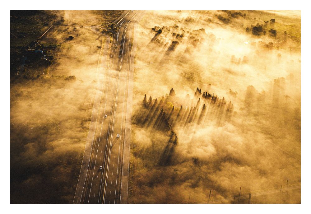A dual carriageway seen from high above. The ground has a thick layer of mist and the sun is very low in the sky creating very long shadows from the surrounding trees in a warm morning light.