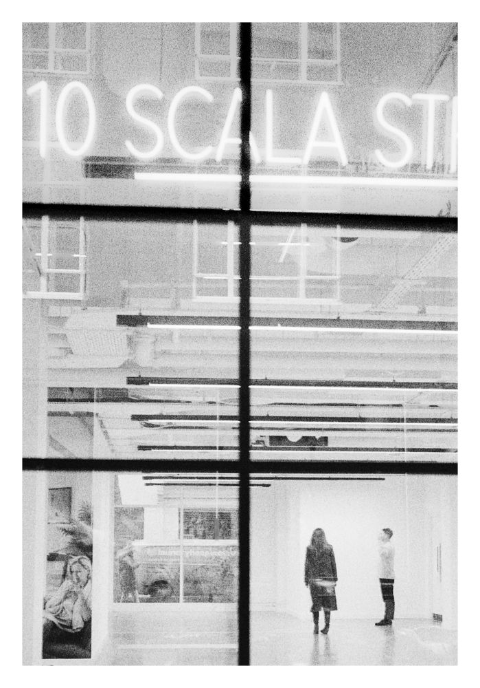 Looking through a six-pane black frame window into an a large white room lit by strip lamps. Two people are framed by the bottom right pane. A work of art is visible in the bottom left pane. A neon sign with the text ‘10 SCALA ST’ is behind the top two frames.