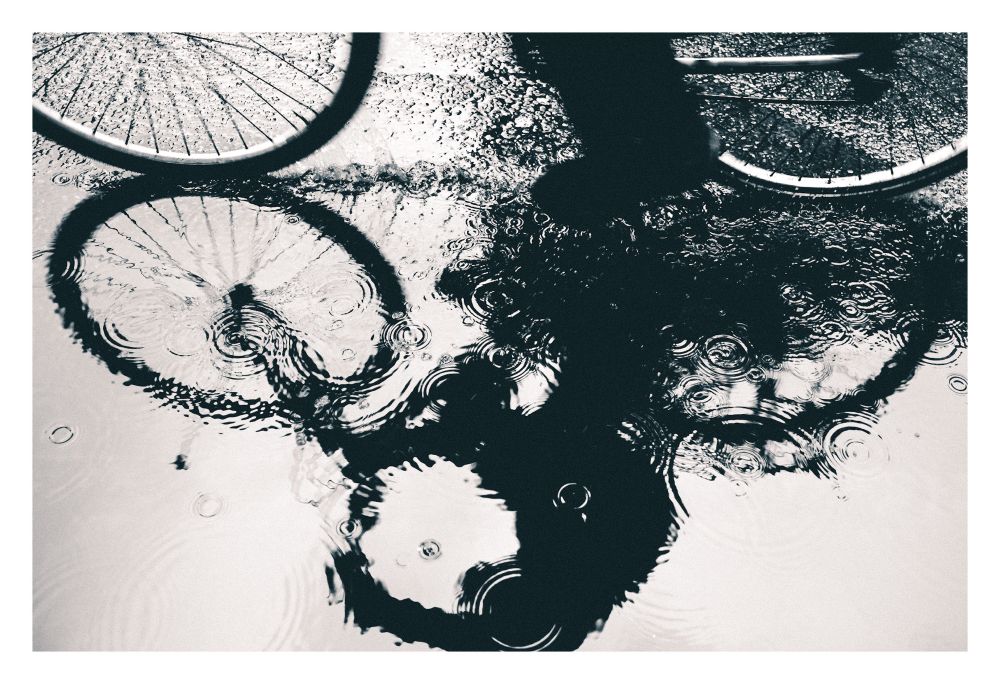 Black and white reflection of a cyclist seen in a puddle with ripples from raindrops.