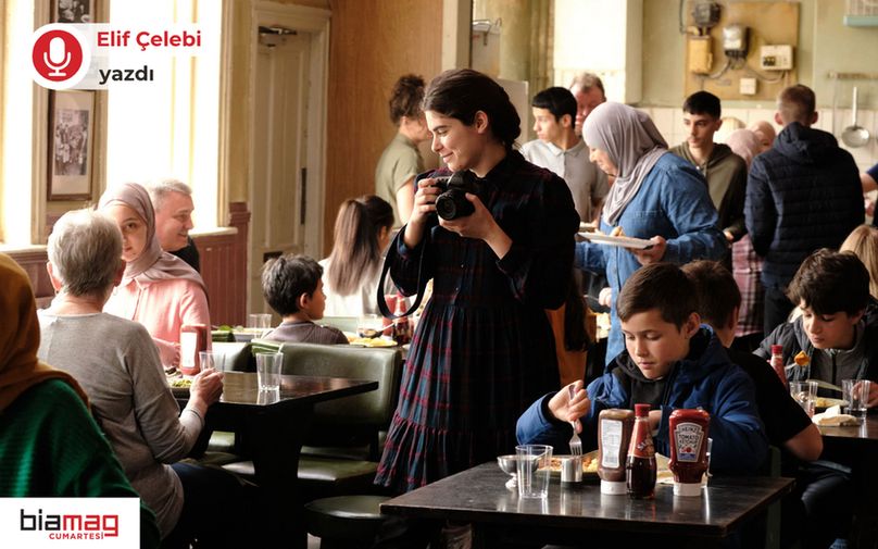 Görselde kalabalık bir yemekhanede insanlar yemek yerken ve sohbet ederken görülüyor. Ortada, elinde fotoğraf makinesiyle gülümseyen genç bir kadın dikkat çekiyor. Farklı yaş gruplarından ve kültürel arka planlardan insanların bir arada olduğu sıcak ve samimi bir ortam yansıtılıyor. Sol üstte “Elif Çelebi yazdı”, sol altta ise “biamag cumartesi” logosu yer alıyor. 