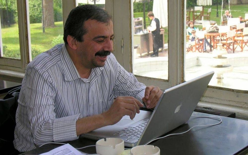 Görselde, cam kenarında bir kafede oturan bir adam görülüyor. Üzerinde açık renkli, çizgili bir gömlek var. Masada bir dizüstü bilgisayar (Apple marka, gümüş renkli), iki beyaz fincan ve bazı belgeler bulunuyor. Adam bilgisayara bakarak gülümsüyor; görünüşe göre bir görüntülü görüşme yapıyor veya bir şey izliyor. Arka planda, camların ardından dış mekânda oturan insanlar, çocuklar ve bir garson dikkat çekiyor. Ortam rahat, doğal ışıkla aydınlanmış ve huzurlu bir atmosfer yansıtıyor.