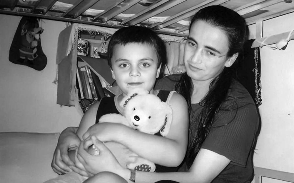 Bu siyah-beyaz fotoğrafta, küçük bir çocuk ve bir kadın birlikte görüntülenmiş. Görselin samimi ve duygusal bir havası var.
Çocuk, kucağında sevimli bir peluş ayı tutuyor ve doğrudan kameraya bakıyor. Yüzünde sakin ve masum bir ifade var.
Kadın, çocuğu arkadan kucaklamış; yüzünde hafifçe gülümseyen ama düşünceli bir ifade taşıyor. Uzun siyah saçları yana taranmış.
İkili, muhtemelen bir ranzanın alt katında oturuyor. Arka planda, kitaplar, asılı bez parçaları ve bir Noel çorabı benzeri süsleme yer alıyor.
Ortam sade ve gösterişsiz; ev içi çekim olduğu anlaşılıyor. Arka plan detayları, mekânın oldukça mütevazı koşullara sahip olduğunu düşündürüyor.
Fotoğraf, güçlü bir anne-çocuk bağı, sevgi ve aynı zamanda yoksulluk ya da zor yaşam koşullarına dair bir hikâye çağrıştırıyor. Siyah-beyaz olması, görüntüye nostaljik ve hüzünlü bir ton katıyor.