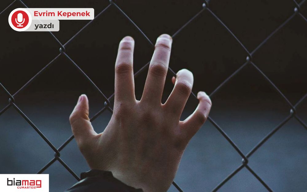 Görselde, parmaklık şeklindeki bir tel örgüye içeriden uzanmış bir el yer alıyor. Arka plan karanlık ve belirsiz; bu da görsele hüzünlü, sıkışmış ya da mahkûmiyet hissi veren bir atmosfer katıyor. Sol üstte “Evrim Kepenek yazdı”, sol altta ise “biamag cumartesi” logosu bulunuyor. Görsel, özgürlük, sınırlar veya tutsaklık temalarını çağrıştırıyor.