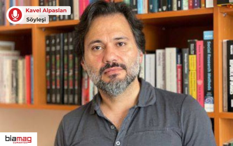 Bu görselde, kitaplık önünde duran bir kişi yer almakta. Kişi, kısa kollu koyu gri bir tişört giymiş ve kameraya doğrudan bakıyor. Hafif dalgalı, orta uzunlukta koyu saçları ve sakallı bir yüzü var. Arka planda, birçok kitabın sıralandığı ahşap raflı bir kitaplık bulunuyor. Kitapların sırt kısımlarında farklı dillerde başlıklar görülebiliyor; bu da kişinin entelektüel bir ortamda bulunduğunu gösteriyor. 
Görselin sol üst köşesinde kırmızı mikrofon simgesiyle birlikte “Kavel Alpaslan - Söyleşi” yazısı yer alıyor. Sol alt köşede ise "biamag cumartesi" logosu bulunmakta. Bu, görselin bir röportaj veya yazı dizisi kapsamında kullanıldığını ve Kavel Alpaslan’ın konu edildiğini işaret ediyor. Genel atmosfer ciddi ve düşünsel bir içeriğe işaret ediyor. 
