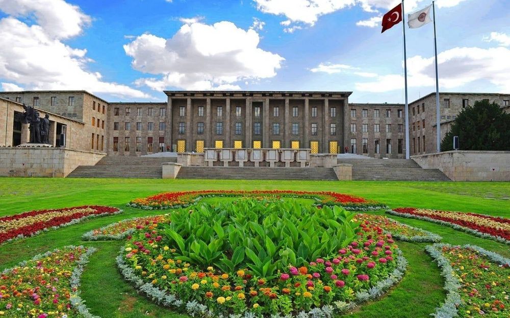 Türkiye Büyük Millet Meclisi (TBMM) binasının ön cephesi, yemyeşil çimlerle kaplı bahçesi ve rengarenk çiçek düzenlemeleriyle birlikte, arka planda Türkiye ve TBMM bayrakları göndere çekilmiş şekilde görülüyor. 