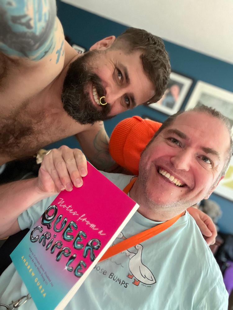 Andrew & Jon.  Jon is a white, shirtless bearded man smiling.  Andrew is a wheelchair using white man holding his book. 