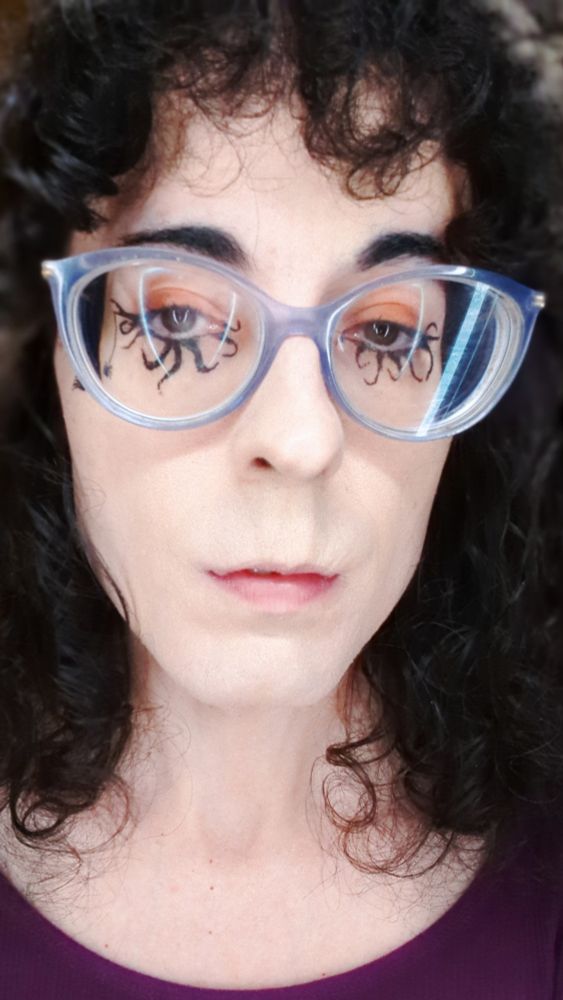 A white woman with curly black hair and bangs. She's wearing purple cat-eye glasses, a fuschia top, orange eyeshadow, and black eyeliner that looks like tentacles extending from her bottom eyelids. There's a little spider dangling from the end of her right eye. She's fully facing the camera and looking down at it slightly, as though in disdain of your pathetic mortal soul.