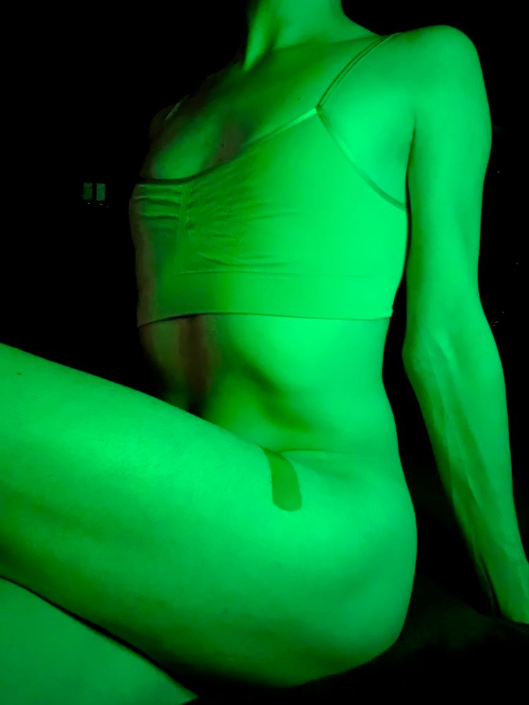 A woman's body from the neck down to the mid-thigh as she basks in just a skin-colored bralette in radioactive green light. She sits with her left side closer to the camera, her left leg bent up to highlight her thighs, and she leans back to reveal the time muscles of her stomach and the swell of her breasts under her tight bralette. Her left thigh bears a bandage from her most recent shot.