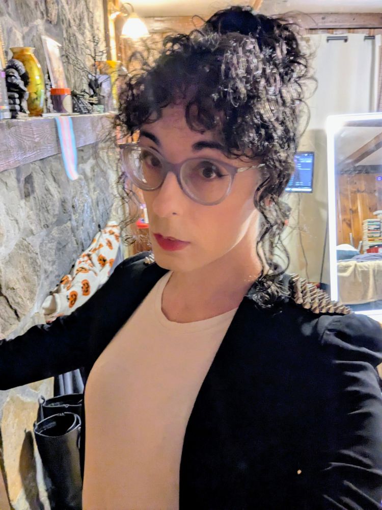 A closeup of a white woman with her curly black hair in a high ponytail with bangs. She wears purple cat-eye glasses, smoky eyeshadow, and red lipstick on her face and her outfit consists of a tight white T-shirt under a black jacket with spiked shoulders.