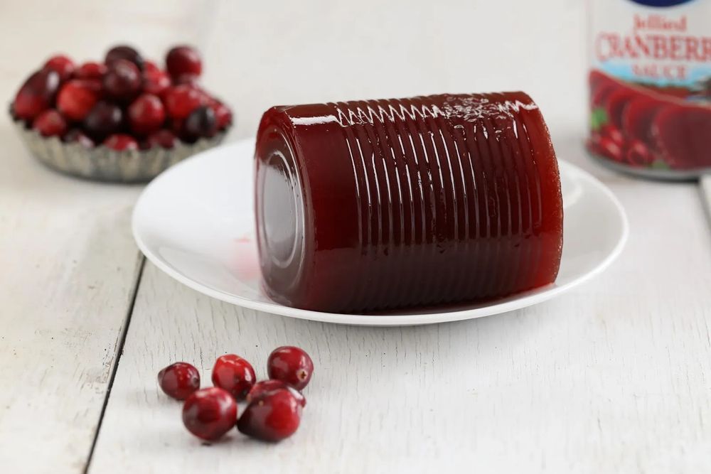 Cranberry sauce from a can in a white dish, on a white table surrounded by actual cranberries and the can in the background.