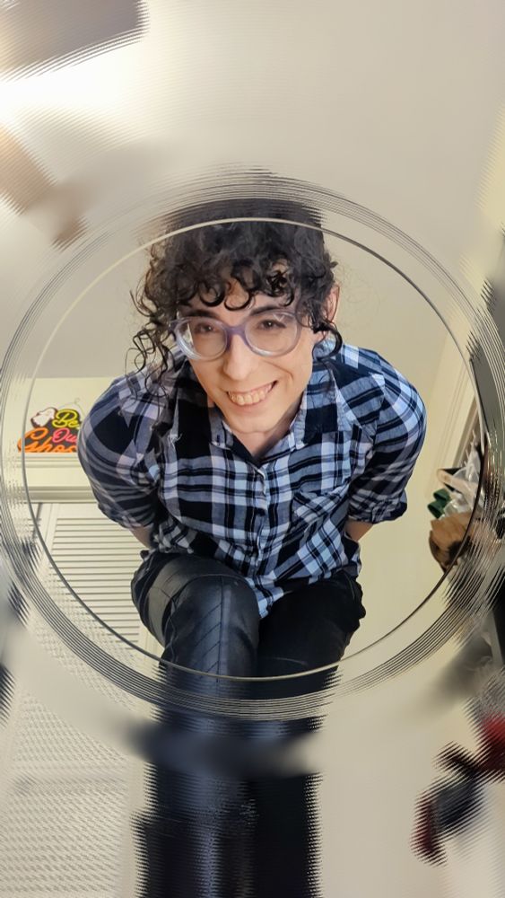 A white woman with curly black hair and bangs is leaning forward and looking down with an amused smirk. She's wearing a black and white flannel, black leather pants, and black calf-high boots. The viewer is looking up at her through the open lid of a clear glass jar.