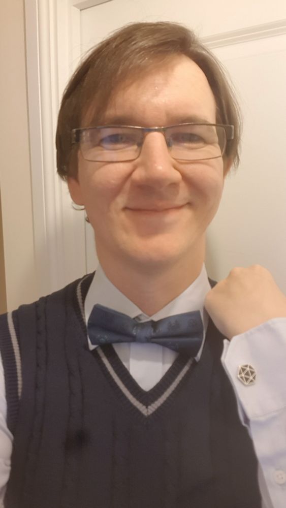 Selfie, where I have a pale blue shirt and navy sweater vest. I have a navy bow tie, with a mix of D&D beholder and the outline of a D20 on it. I am also showing a cufflinks of a D20, with silver trim and filled in navy. The shot is from the upper torso and up.