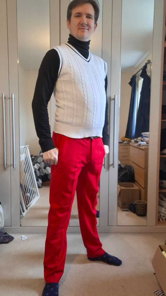 Selfie of me in bright red smart trousers, with a brown belt, and a black turtleneck under a white v-neck sweater vest.