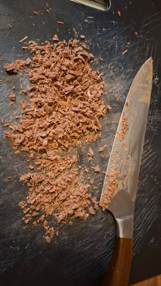 A black chopping board, with a long chef's knife beside some finely chopped chocolate 