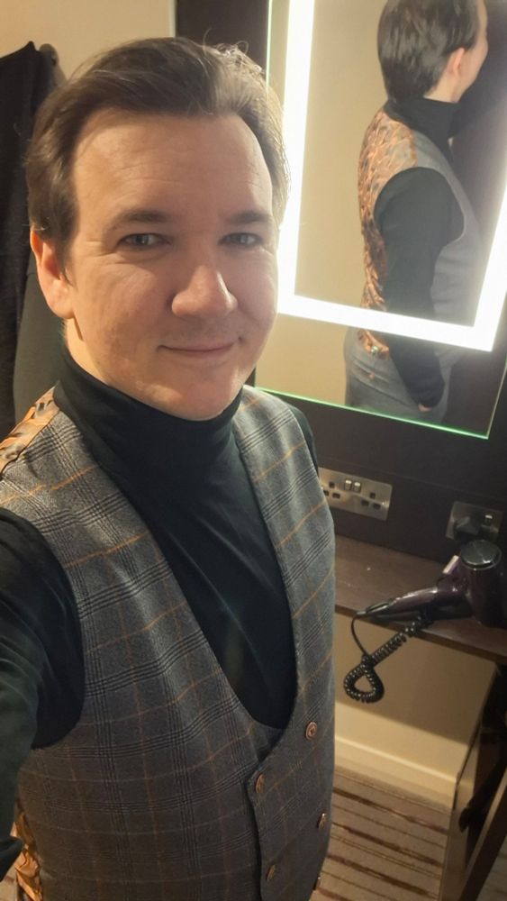 Selfie of white man in black turtleneck and double-breasted grey waistcoat, with hiseeflection behind him in a mirror.