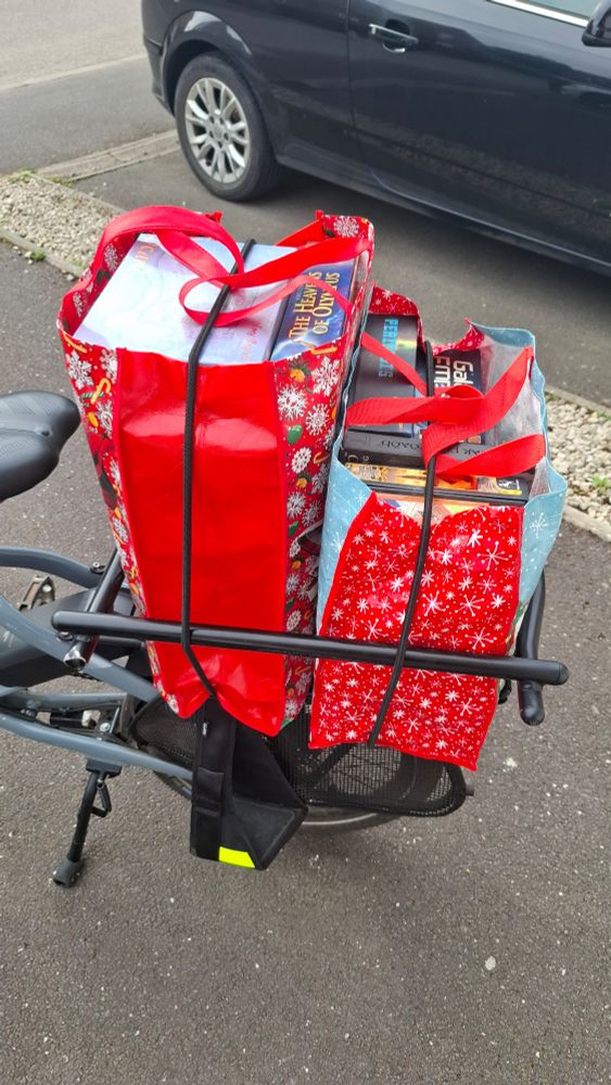 Back of a cargo bike, with two large red bags with snowflakes on. Inside are multiple boxes of board games. 