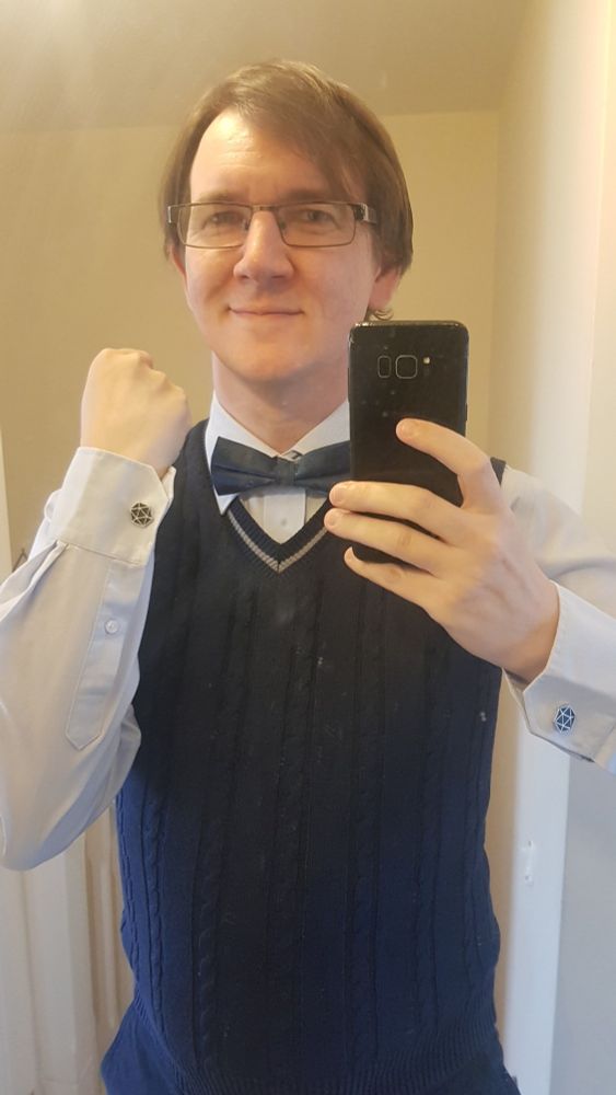 Selfie, where I have a pale blue shirt and navy sweater vest. I have a navy bow tie, with a mix of D&D beholder and the outline of a D20 on it. I am also showing a cufflinks of a D20, with silver trim and filled in navy. The shot is from the torso and up, reflected in a mirror.