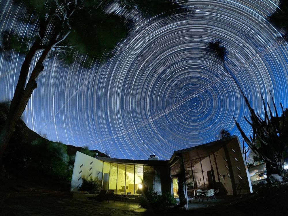 Whirl of hundreds of time-lapsed stars in the night sky over a softly lit modernist house between a pine and a palm tree. 