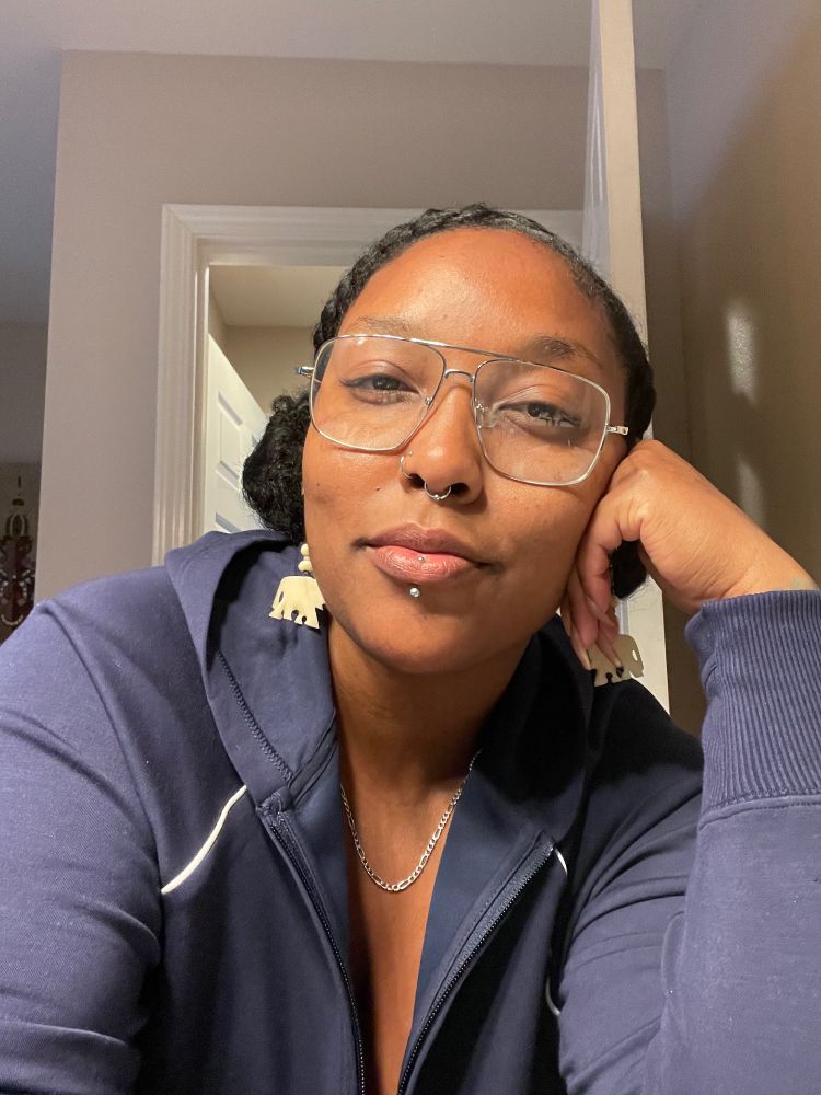 A Black woman with facial piercings wearing glasses and a blue long sleeve smiles at the camera in a selfie. She has her hair in two braids pulled into buns and is wearing elephant earrings.