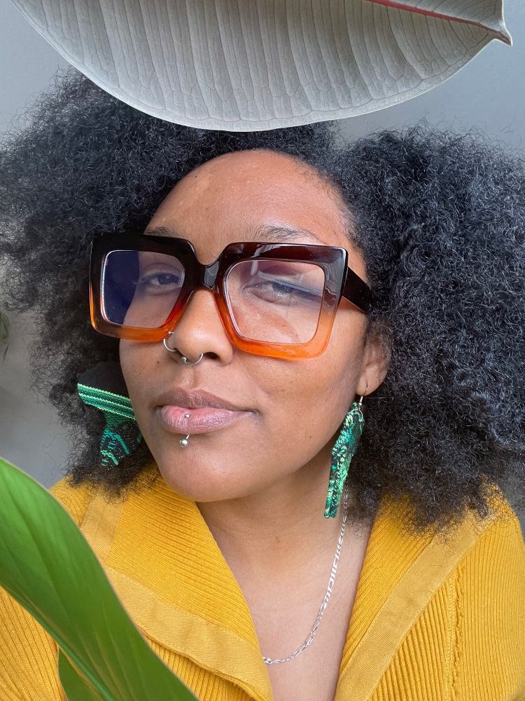 A Black woman with an Afro wearing a yellow top and ombré glasses smirks at the camera through two houseplant leaves.
