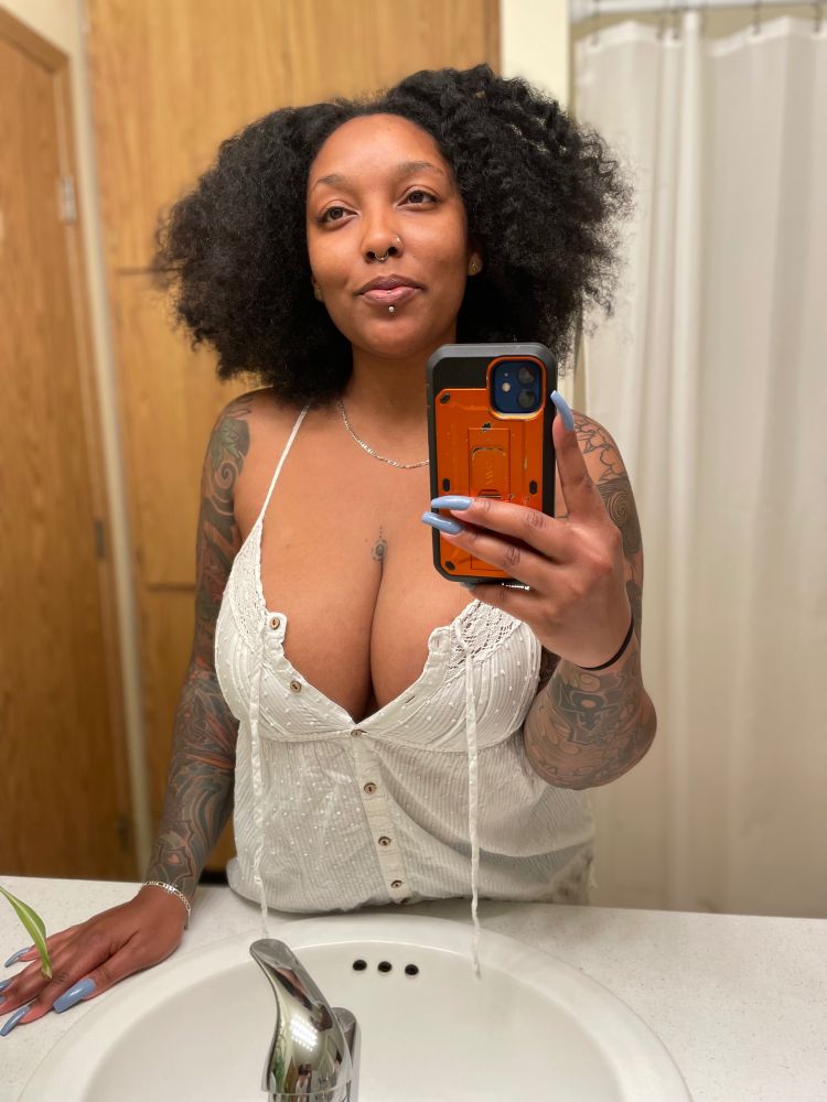 A Black woman with arm tattoos and a wavy afro wearing a white button down linen tank top takes a selfie in a mirror. 