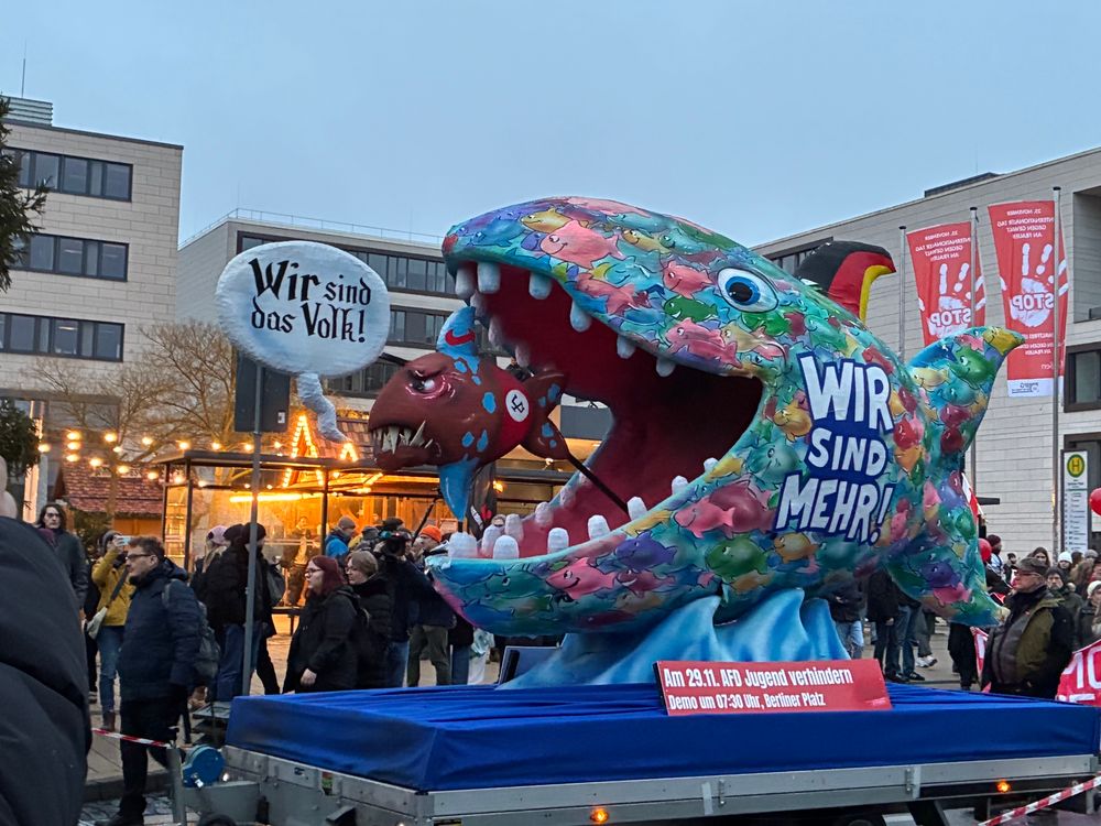 Demo gegen die Neugründung der A*D-Jugendorganisation am Berliner Platz in Gießen heute früh. Man sieht einen Aufbauwagen wo ein Wir sind das Volk-Fisch mit rot schwarz weißer Binde von einem größeren Wir sind mehr-Fisch gefressen wird. Auf dem großen sind sehr viele bunte Fische gemalt. 