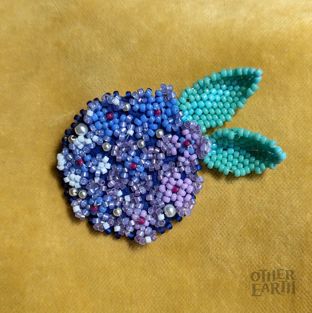 A brooch made out of little four-petal flowers in shades of periwinkle, violet, and lilac stitched on a cobalt blue background. Two large leaves in shades of turquoise stick out from the brooch body. 