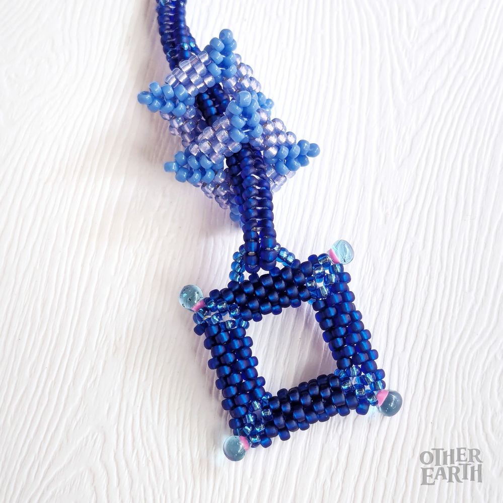 A close up of a bracelet in shades of blue lies upon a white background. The body of the bracelet is a rich sapphire blue with periwinkle peyote squares threaded through it. The toggle clasp is also made from a sapphire peyote square studded with blue and pink drop beads along each edge. 