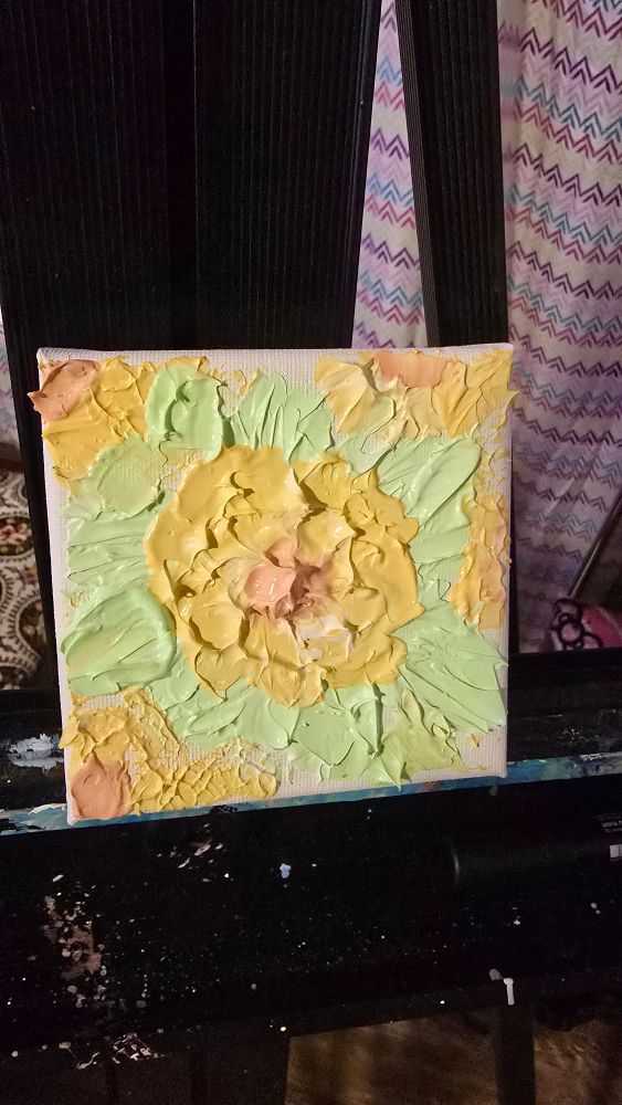 Photo of a small paining. The center is a yellow flower with an orange center with light grean leaves around it and othe flowers on the edges. It is sitting on a black easel with a multicored shed behind it. The painting has some three dimensionionality to it with the pettals coming to a point off of the canvas. The leaves around the flower also have some mild 3 dimensionality to them although are a bit flat and lacking some shape. 