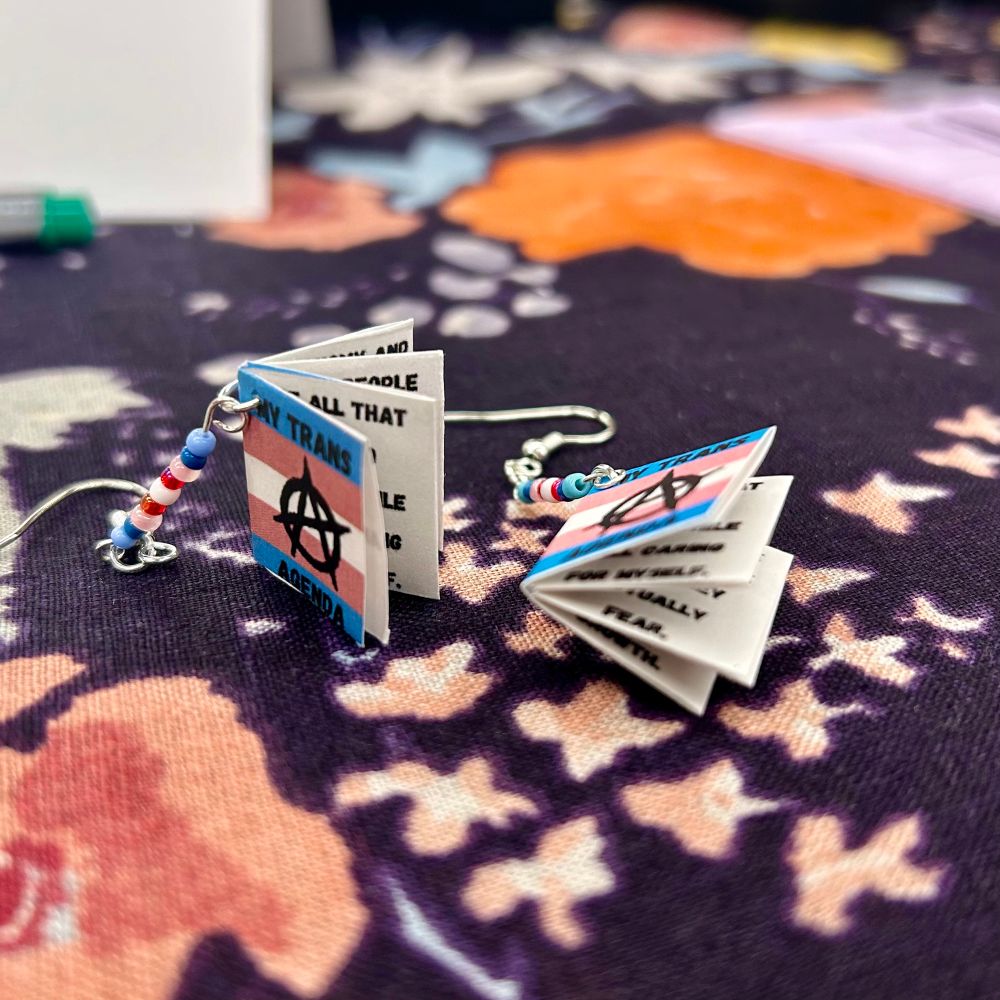 A pair of tiny zines titled “MY TRANS AGENDA” are affixed to earrings and sitting decoratively on a patterned cloth.