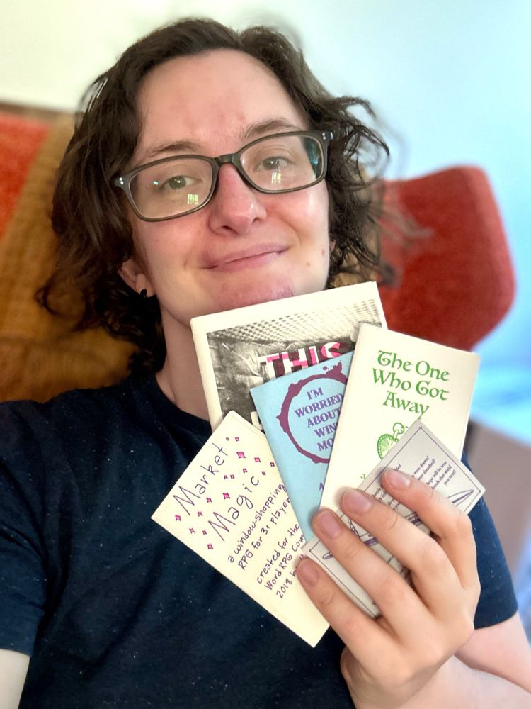 A white, brunette transmasc person smiles while holding up a large handful of colorful risogrpah printed zines next to their face. 
