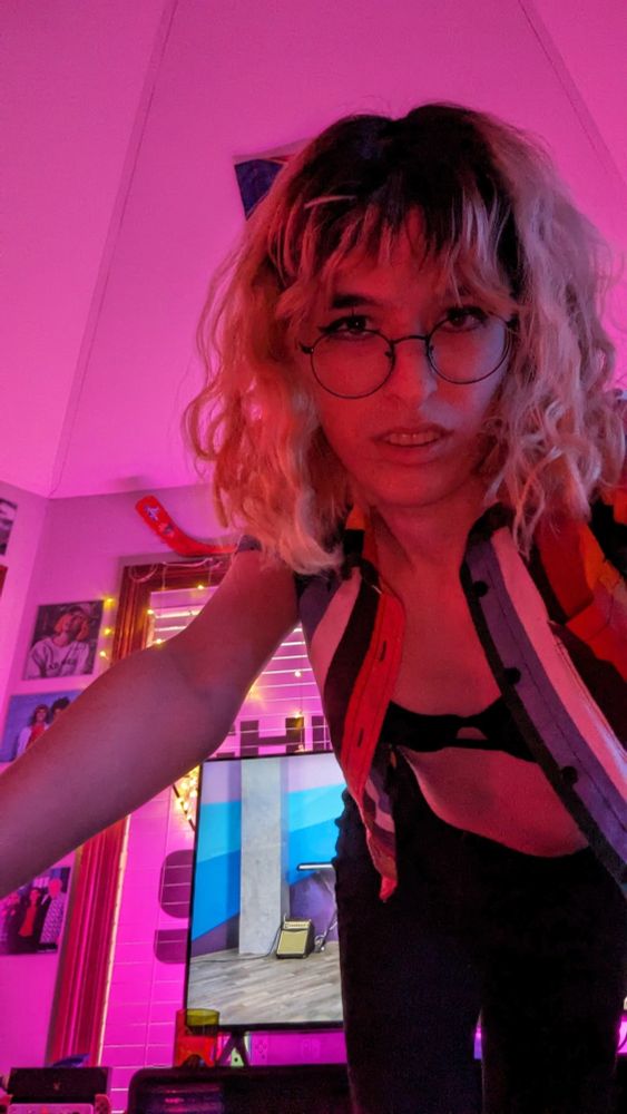 juniper bending over the camera in pink lighting. cropped button-up that's open to reveal just a bra. black jeans. she is hot