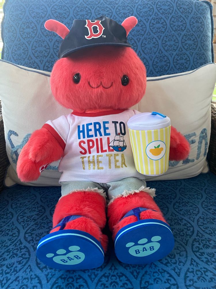 A red anthropomorphic lobster plush dressed in a white T-shirt with red collar and cuffs that says Here to Spill the Tea in red, blue, and glittery gold. The T-shirt has a sketch of a Colonial style shop with the Build-À-Bear logo on it. The lobster is also wearing light blue frayed denim shorts, and darker blue sandals. They are holding a plush lemonade in a cup with a straw sticking out of it. Atop their head is a navy blue Red Sox cap. They are sitting on a wicker chair with blue upholstery and a white toss pillow behind them.