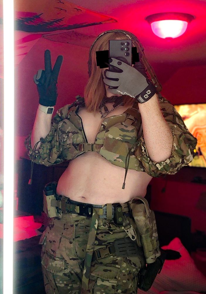 a pinup girl, langley, poses for a propaganda photo in a multicam bikini top and combat gear. she makes a V with her fingers