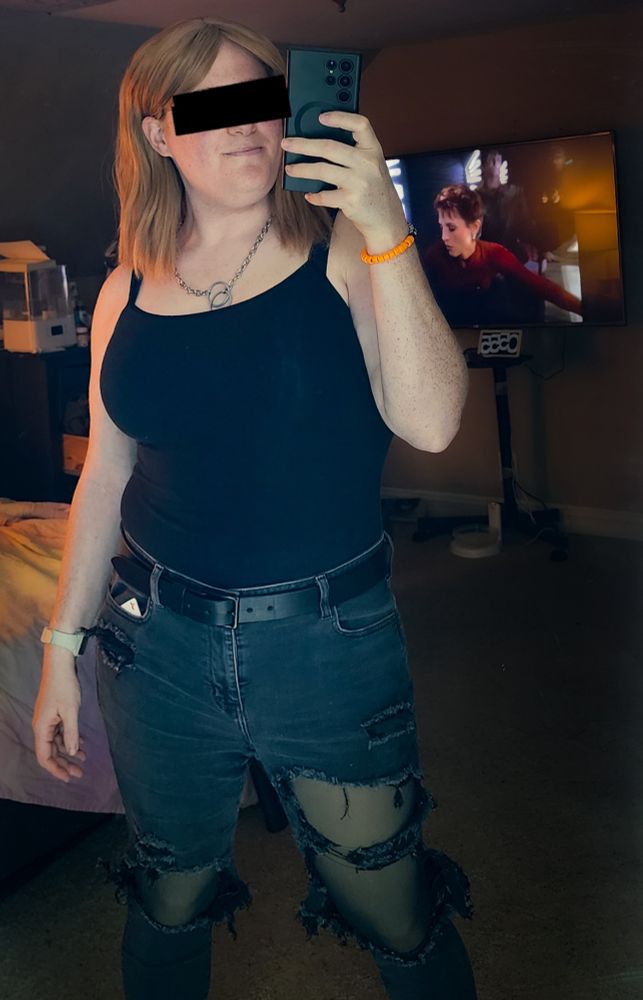 a beautiful girl with strawberry blond hair, langley, stands in front of a mirror Ina confident pose. she's wearing a black tank top and ripped black jeans with leggings. she's very hot