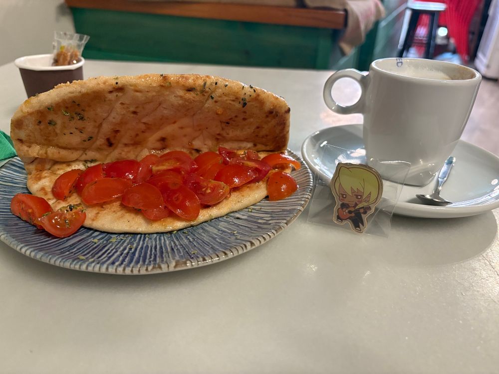 A la izquierda tostada de pan pita griego con tomate y aceite de oliva y a la derecha café capuccino y en el platito del café está el pin de Klavier Gavin (Ace Attorney)