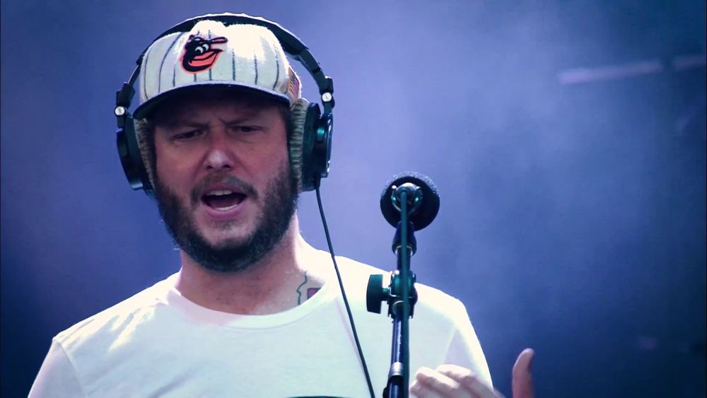 Justin Vernon / Bon Iver wearing an Orioles hat under headphones. He is standing in front of a microphone.