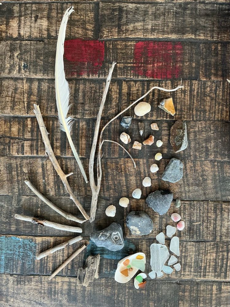 auf einem tisch (auf altes holz getrimmt, dunkler mit roten & türkisen farbresten) liegt, was ich am strand gesammelt habe: kleine schwemmhölzchen, steine, muscheln und eine weiße feder. und sehr kleine bunte bis etwas größere weiße abgeschliffene glasscherben. die mag ich am liebsten.